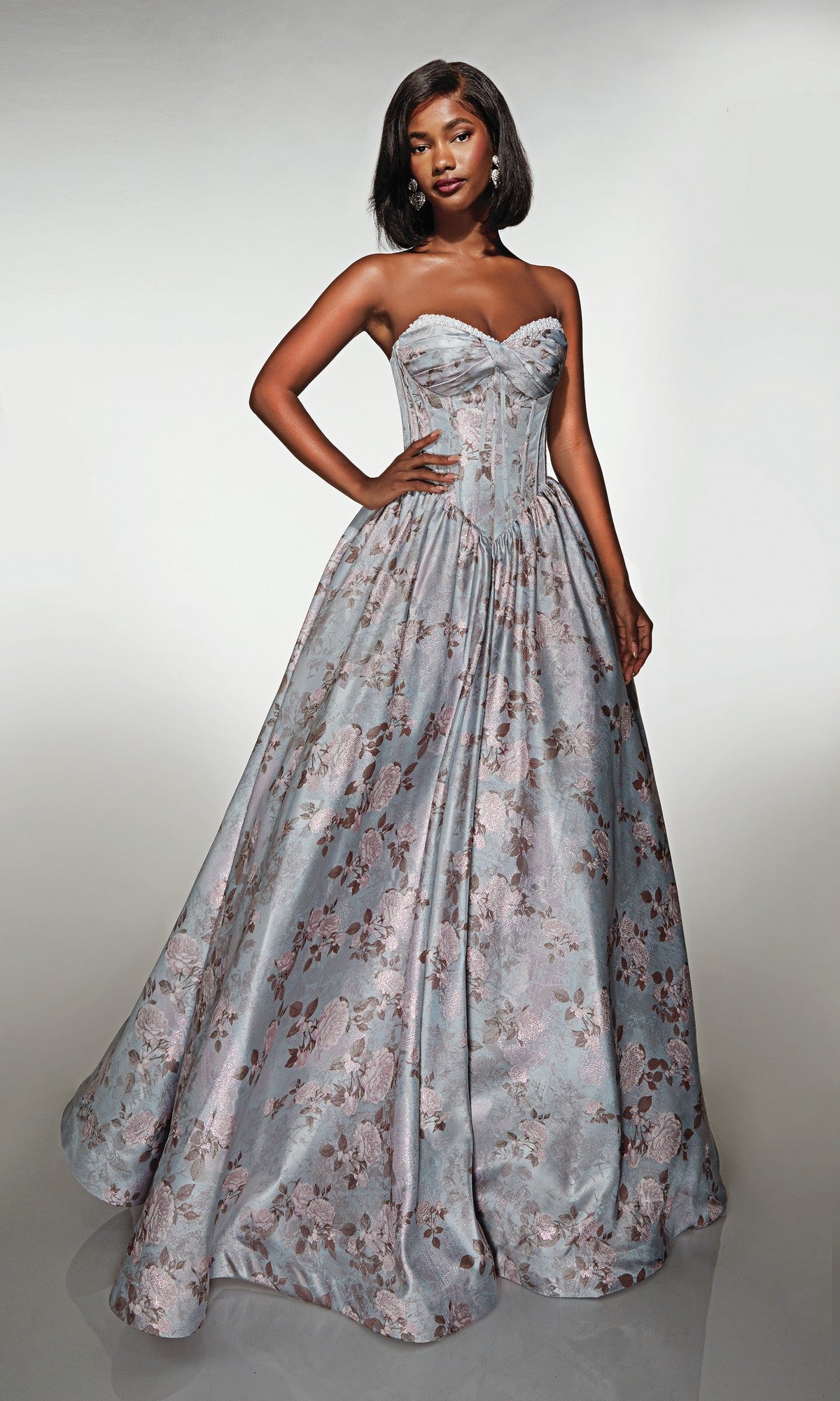 A woman stands in the Alyce 62181 long prom dress, featuring a strapless pink/jade ball gown with a fitted lace-up corset bodice and floral print, set against a plain background.